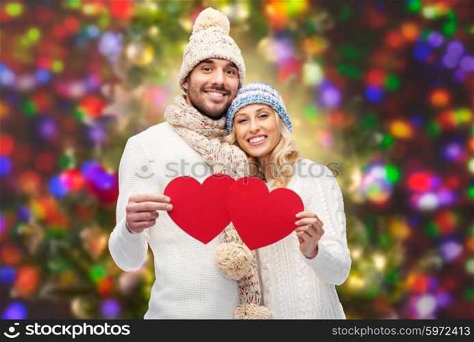 love, valentines day, couple, christmas and people concept - smiling man and woman in winter hats and scarf holding red paper heart shapes over holidays lights background