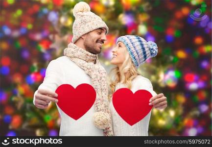 love, valentines day, couple, christmas and people concept - smiling man and woman in winter hats and scarf holding red paper heart shapes over holidays lights background