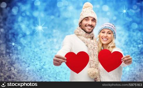 love, valentines day, couple, christmas and people concept - smiling man and woman in winter hats and scarf holding red paper heart shapes over blue glitter or lights background
