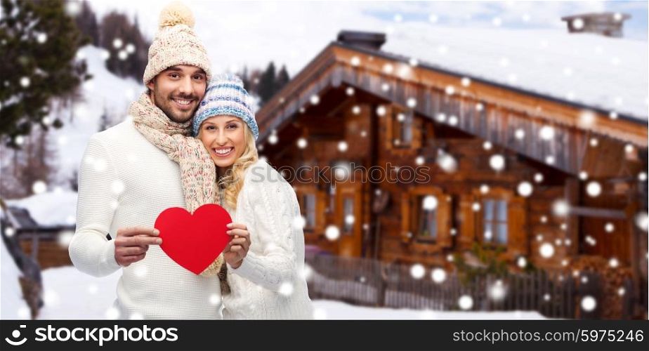 love, valentines day, couple, christmas and people concept - smiling man and woman in winter hats and scarf holding red paper heart shape over wooden country house and snow background