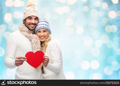 love, valentines day, couple, christmas and people concept - smiling man and woman in winter hats and scarf holding red paper heart shape over blue holidays lights background