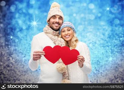 love, valentines day, couple, christmas and people concept - smiling man and woman in winter hats and scarf holding red paper heart shapes over blue glitter and holidays lights background