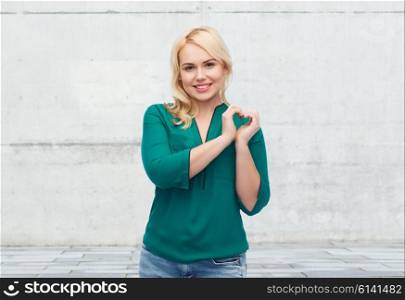 love, valentines day, gesture, plus size and people concept - smiling young woman in shirt and jeans showing heart shape hand sign over gray concrete wall background