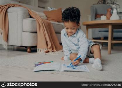 Lovely african child sitting on floor at home in stylish living room with colored felt tip pens and drawing in album with focused expression on his face while spending leisure time at home. Lovely african child sitting on floor and drawing while playing at home