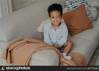 Lovely afro american child with curly hair and cute smile sitting on sofa with beige color elements pillows and coverlid, looking up on camera and smiling while playing in living room at home. Lovely Afro American child with curly hair and cute smile looking up on camera and smiling
