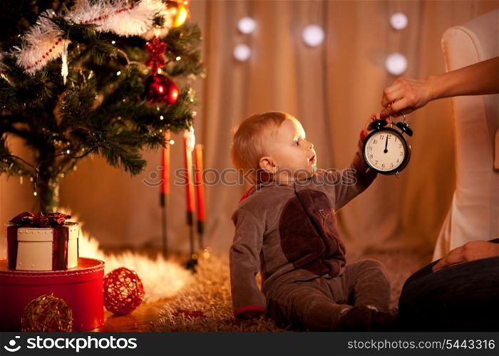 Lovely baby near Christmas tree holding with mother alarm clock&#xA;