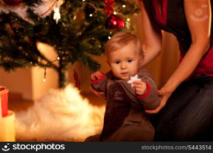 Lovely baby sitting with mama near Christmas tree and looking on Christmas toy&#xA;