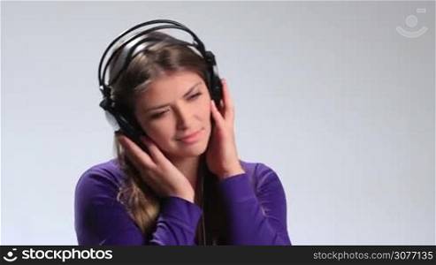 Lovely brunette girl with eyes closed holding both hands on headphones and enjoying beautiful song on mp3 player. Charming young woman opening her eyes, looking at the camera enigmatically and sending air kiss while listening music in earphones.