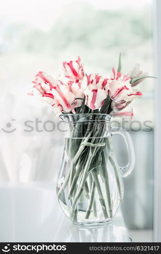 Lovely bunch of tulips in glass jug on white table at window. Flowers in interior design. Cozy home. Springtime
