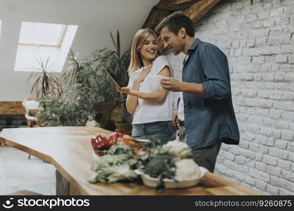 Lovely cheerful young couple cooking dinner together, looking recipe at digital tablet and having fun at rustic kitchen