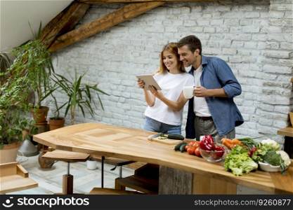 Lovely cheerful young couple cooking dinner together, looking recipe at digital tablet and having fun at rustic kitchen
