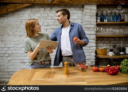 Lovely cheerful young couple cooking dinner together, looking recipe at digital tablet and having fun at the rustic kitchen