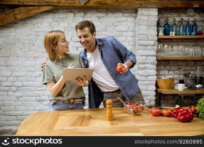 Lovely cheerful young couple cooking dinner together, looking recipe at digital tablet and having fun at the rustic kitchen