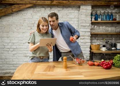 Lovely cheerful young couple cooking dinner together, looking recipe at digital tablet and having fun at the rustic kitchen