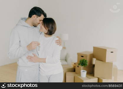 Lovely couple embrace and feel happy, holds keys from new modern first house, move in apartment for living together, pose in empty room with packed carton boxes, feel love to each other. Real estate