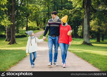 Lovely couple embraces each other, looks at their small good looking daughter, walks across green park. Little adorable girl keeps father`s hand, has pleasant conversation with her parents