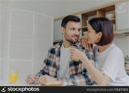 Lovely couple look at each other with love, woman holds glass of juice, pose together near kitchen table, have pleasant talk during breakfast time, enjoy domestic atmosphere. Happy family concept