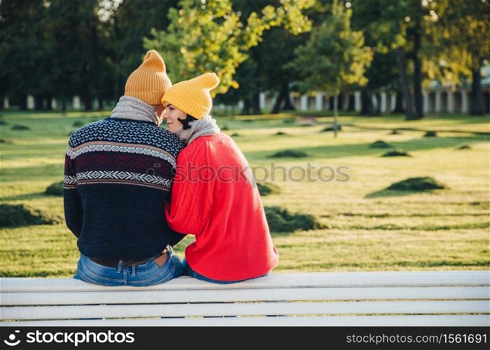 Lovely couple sit on bench together, wear warm clothes and knitted hats, embrace each other, express love and good relationship, enjoy beautiful landscapes and fresh air in park. Relationship concept