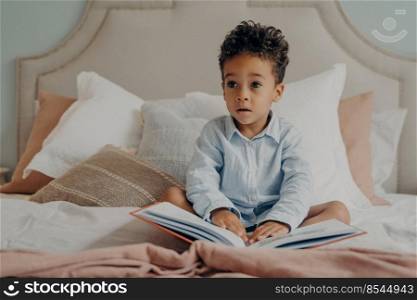Lovely curly haired afro american little boy kid in casual clothes sitting on bed full of pillows with story book on his lap at home, looking aside with curious face expression. Child playing indoors. Cute curly haired afro american little boy sitting on bed with book