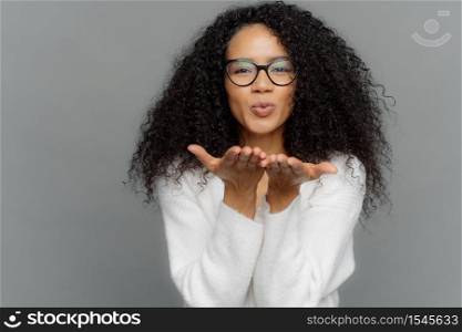 Lovely curly woman sends air kiss, has lips folded, keeps palms outstretched, wears transparent glasses, white jumper, expresses affection to boyfriend, isolated on grey background. Mwah to you