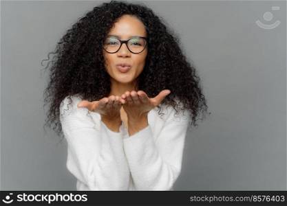 Lovely curly woman sends air kiss, has lips folded, keeps palms outstretched, wears transparent glasses, white jumper, expresses affection to boyfriend, isolated on grey background. Mwah to you
