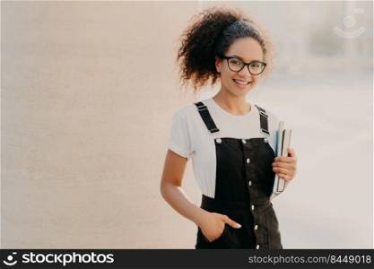 Lovely curly woman with combed hair, dressed in white t shirt, sarafan, keeps hand in pocket, holds book and textbook, wears optical glasses, poses at unversity c&us, smiles happily at camera