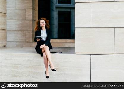 Lovely curly woman with confident look, sitting alone, demonstrating her slender long legs, dressed in black jacket with skirt, holding tablet, looking schedules or diagrams, making business report
