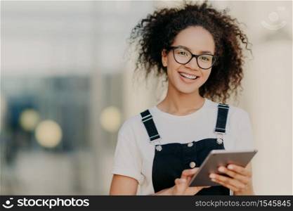 Lovely dark skinned ethnic woman with crisp hair combed in pony tail, smiles gently, wears optical glasses, white t shirt and overalls, holds modern touchpad, reads book online, chats with friends