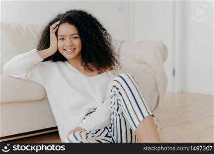 Lovely dark skinned woman with crisp hair, sits on floor near sofa, wears white sweater and striped trousers, enjoys positive emotions, enjoys weekend at home. People, rest, happiness concept