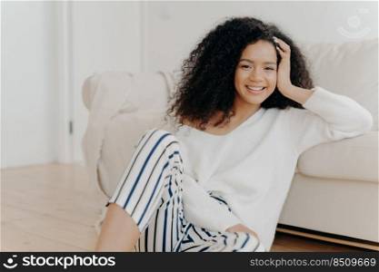 Lovely dark skinned woman with crisp hair, sits on floor near sofa, wears white sweater and striped trousers, enjoys positive emotions, enjoys weekend at home. People, rest, happiness concept