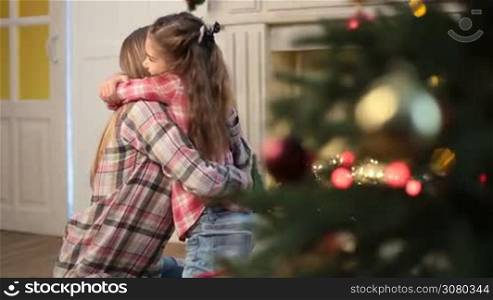 Lovely daughter with long brown hair embracing and caressing her affectionate mother while family relaxing on Christmas eve at home. Little cute girl expressing her love and affection to mom during winter holidays over xmas decorated room background.