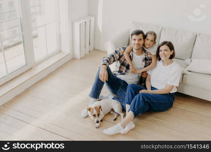 Lovely European family of father, mother and their daughter sit on floor near sofa in spacious white room, pedigree dog lies near, have moving day, change place of living, express happiness.