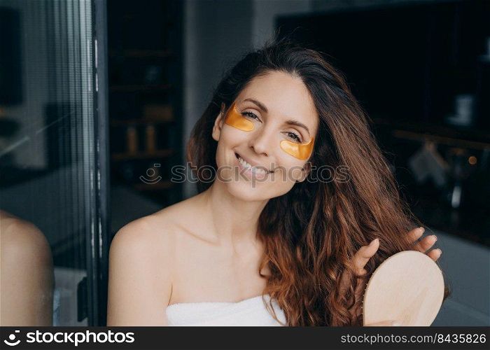 Lovely european woman combs her soft curly hair with hairbrush. Girl applying eye patches after bathing. Young happy woman takes shower at home and doing hair. Weekend morning at home.. Lovely young woman combs her curly hair with hairbrush. Girl applying eye patches after bathing.