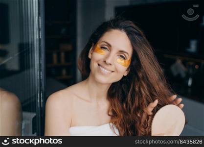 Lovely european woman combs her soft curly hair with hairbrush. Girl applying eye patches after bathing. Young happy woman takes shower at home and doing hair. Weekend morning at home.. Lovely young woman combs her curly hair with hairbrush. Girl applying eye patches after bathing.
