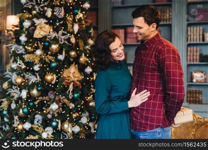Lovely family couple: pretty wife stands near her husband who prepares New Year present for her. Good relationnship concept. Woman and man stand against decorated Christmas tree. Winter holidays