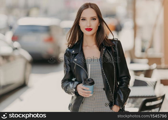 Lovely fashionable woman with makeup, bright red painted lips, wears black leather jacket, strolls at urban setting, stands near cafeteria, holds takeaway coffee, enjoys spare time. People, lifestyle