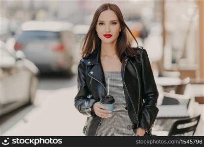 Lovely fashionable woman with makeup, bright red painted lips, wears black leather jacket, strolls at urban setting, stands near cafeteria, holds takeaway coffee, enjoys spare time. People, lifestyle