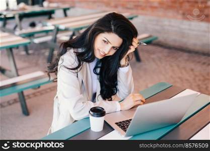 Lovely female businesswoman with dark long hair holding pen in her hand sitting outdoors using laptop computer for her work drinking coffee trying to make project. Adorable brunette female working