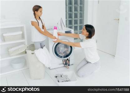 Lovely girl helps mum with washing, stands in basket, gives bottle of detergent or washing powder , woman stands on knees, loads washing machine happy to get help from daughter, puts clothes in washer