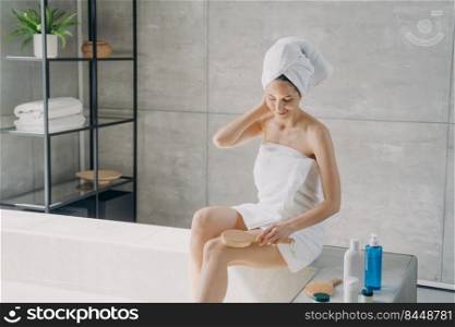 Lovely girl is making anti cellulite body massage with brush in bathroom. Attractive caucasian lady wrapped in towel after bathing. Woman takes shower at home. Morning beauty routine and bodycare.. Lovely caucasian girl is making body massage with brush in bathroom. Morning bodycare routine.