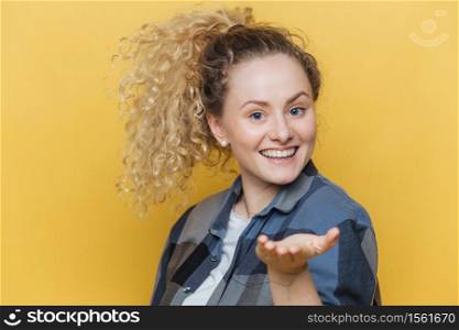 Lovely glad woman stretches hand in foreground, has broad smile, demonstrates perfect white teeth, wears checkered shirt, poses against yellow background. Smiling satisfied female model indoor