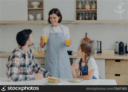 Lovely happy mother wears apron, holds two glasses of orange juice, talks to husband and daugher, prepares breakfast. Three membered family have dinner time at kitchen. Togetherness concept.