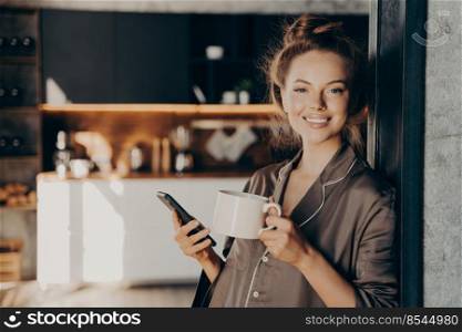 Lovely happy young woman having her morning coffee while checking new emails and notifications on smartphone standing in kitchen broadly smiling after waking up, spending weekend at home. Lovely happy young woman having her morning coffee while checking new emails and notifications on smartphone