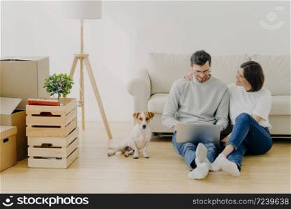 Lovely husband and wife sit in new apartment with laptop computer, discuss home repair project, pose in living room with sofa, stack of boxes and pedigree dog. Moving and relocation concept.