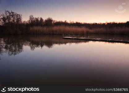 Lovely image of late sunset sky over calm lake landscape with long fishing jetty pier and vibrant colors