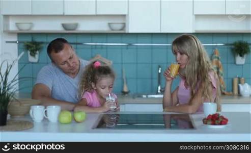 Lovely little curly daughter drinking milk while sitting on father&acute;s lap during family morning breakfast in kitchen. Cheerful family with adorable little girl sharing healthy breakfast together while spending weekend at home.