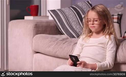 Lovely little girl sitting on the floor near sofa and playing on mobile phone. Smiling child using cellphone at home.