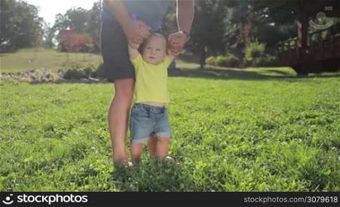 Lovely little toddler baby boy with blue eyes learning to walk barefoot on green grassy lawn with father&acute;s help. Cute infant child taking first steps on grass in summer park while caring dad holding his hands. Slow motion. Steadicam stabilized shot.