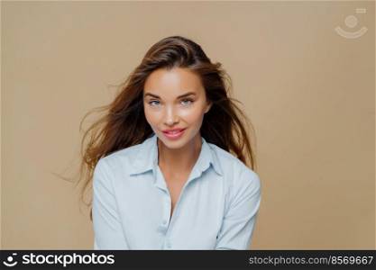 Lovely millennial female looks with satisfied expression at camera, has long hair, dressed in stylish shirt, has natural beauty, models against brown background, wears makeup, has lively talk
