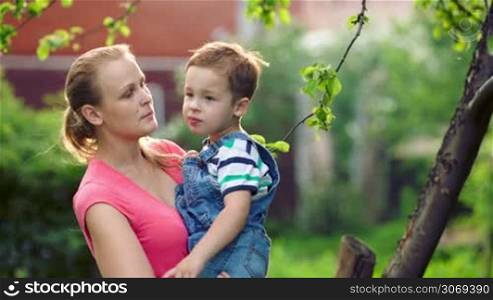 Lovely mother and her little son talking outdoor in the garden. Mom holding her son in arms and kissing him